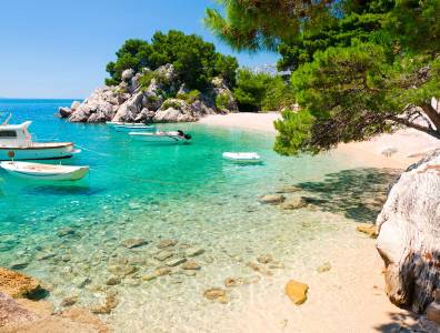 Kleine Boote liegen vor Anker in einer türkisfarbenen Bucht mit Sandstrand, Felsen und Pinien