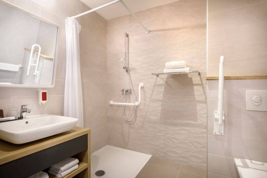 Ein modernes barrierefreies Badezimmer mit beigen Wandfliesen. Es verfügt über eine offene, ebenerdige Dusche mit Haltegriffen, einem höhenverstellbaren Duschkopf und einem Duschvorhang.