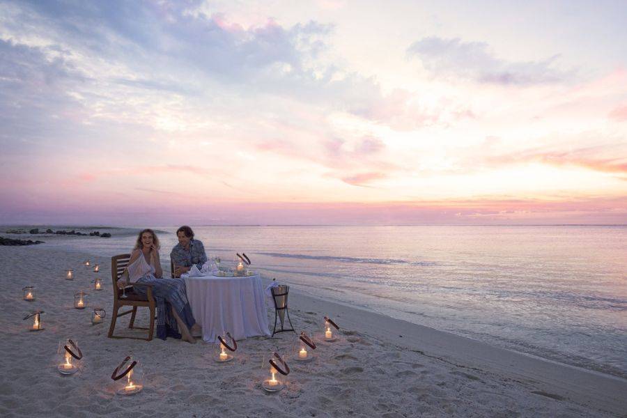 © DINAROBIN BEACHCOMBER Paar beim romantischen Candle-Light-Dinner am Strand bei Sonnenuntergang, mit kleinen Laternen im Sand und ruhigem Meer im Hintergrund.