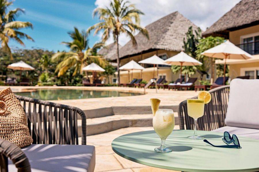 Dieses Bild zeigt eine entspannte Poollandschaft in einem tropischen Resort. Im Vordergrund befinden sich zwei erfrischende Getränke auf einem kleinen Tisch, umgeben von gemütlichen Sitzmöbeln.