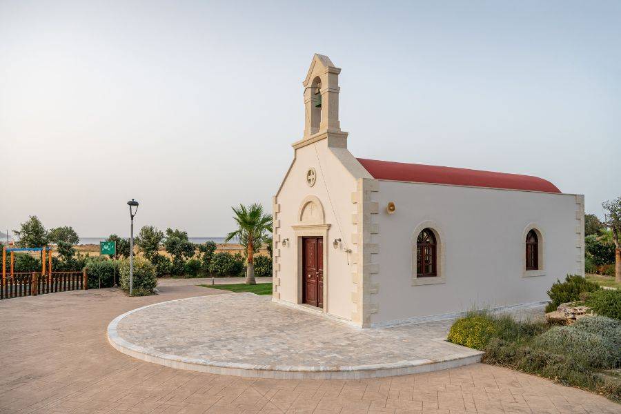 Weiß-beige Kapelle mit rotem Dach und Glockenturm in ruhiger, gepflegter Umgebung, nahe dem Meer gelegen.