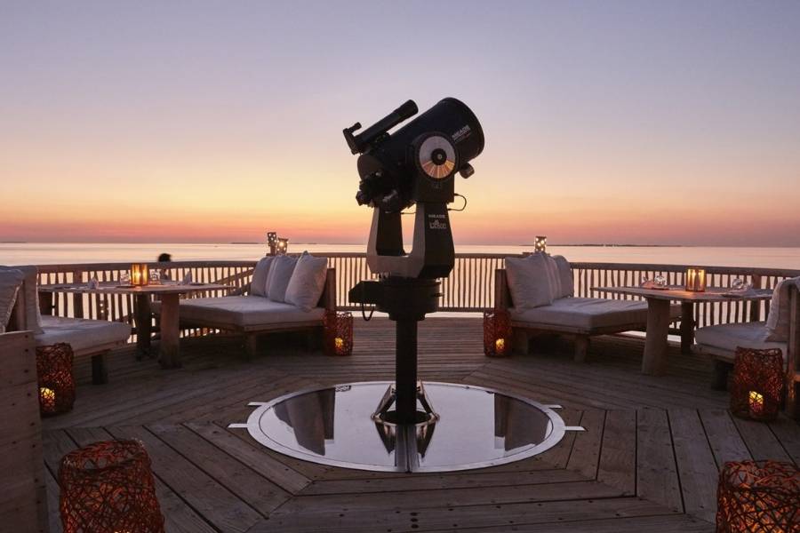 Teleskop auf einer Holzterrasse im Soneva Jani, umgeben von gepolsterten Sitzplätzen und Laternen bei Sonnenuntergang