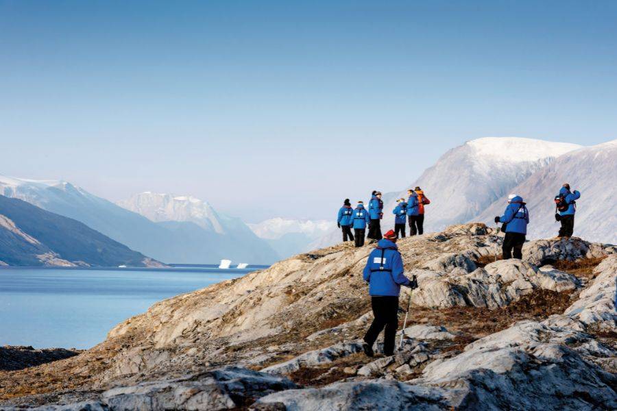 Expeditionsteilnehmer in blauen Jacken wandern entlang einer felsigen Küste in arktischer Landschaft mit Meer und Bergen
