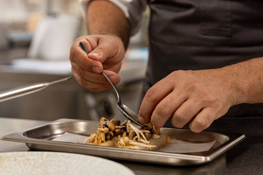 Ein Koch in einer professionellen Küche richtet sorgfältig ein Gericht mit Pilzen auf einem Tablett an.