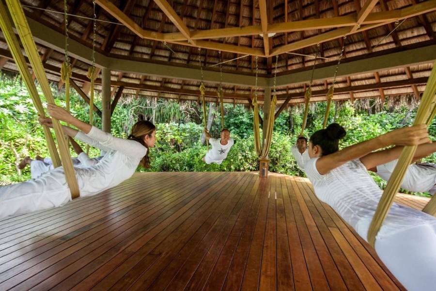 Gruppe beim Aerial Yoga in gelben Tüchern unter dem Strohdach eines offenen, von tropischem Grün umgebenen Pavillions.