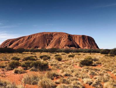 Roter Monolith Uluru im Outback von Australien, umgeben von Buschlandschaft und rotem Sand unter klarem Himmel