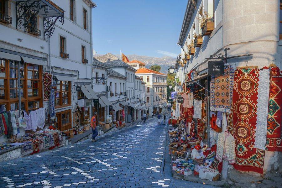 ine malerische Straße mit Kopfsteinpflaster in einer historischen Stadt. Auf beiden Seiten der Straße befinden sich Geschäfte und Stände, die traditionelle Textilien, Teppiche und handgefertigte Waren ausstellen. 