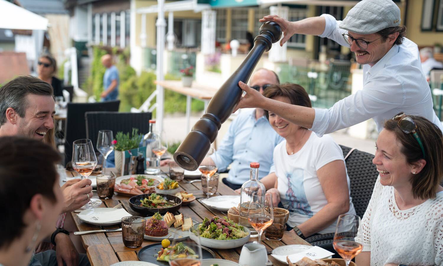 Gesellige Familienfeier bei Paul der Wirt mit Gästen am Tisch & mit Kellner, der mit übergroßer Pfeffermühle Speisen würzt