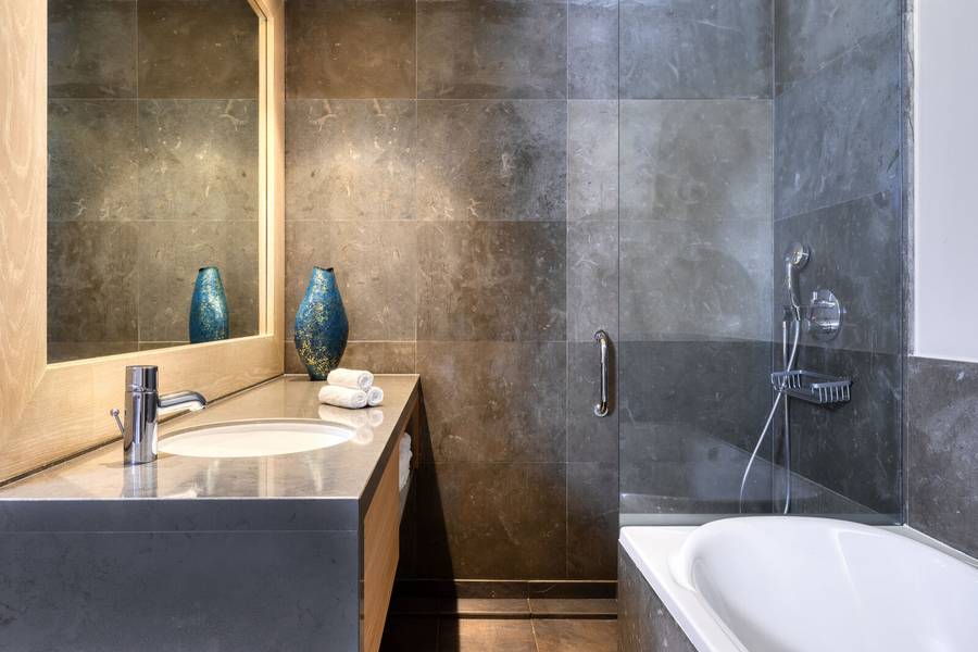 Ein stilvolles Badezimmer mit grauen Steinfliesen, einem großen Spiegel, einer weißen Badewanne und einer modernen Dusche. Eine dekorative blaue Vase setzt einen farblichen Akzent.