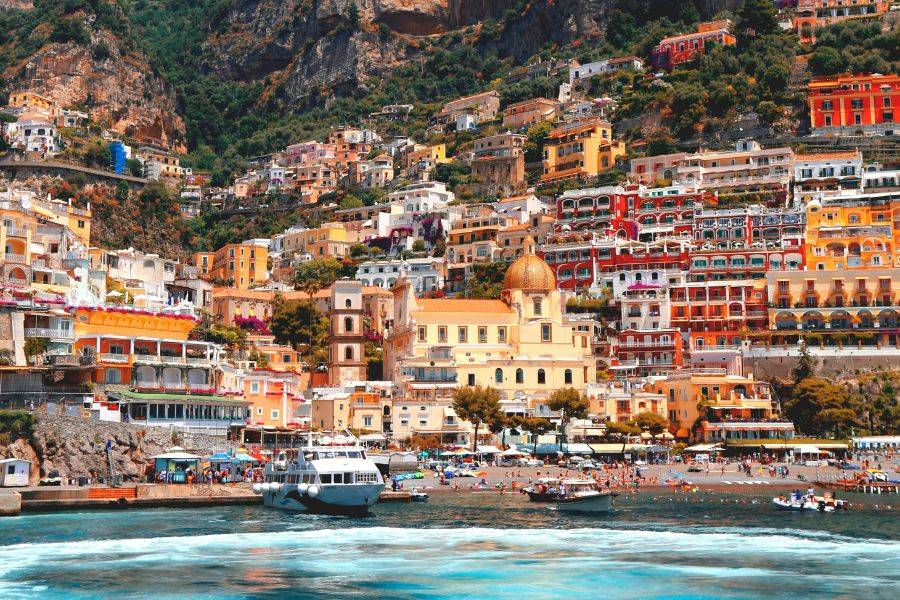 Bunter Küstenort Positano mit terrassenförmig angelegten Häusern an einem steilen Hang, Blick vom Meer auf Strand, Kirche und Boote.