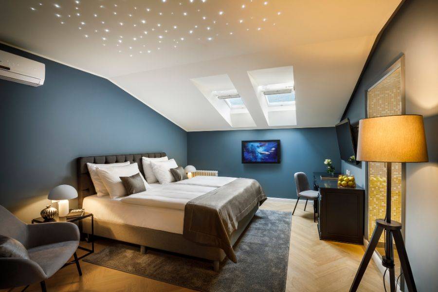 Stilvolles Dachzimmer mit gemütlichem Bett, schräger Decke, Sternenhimmel-Effekt, Oberlichtern und einer eleganten Einrichtung in Blau- und Grautönen.