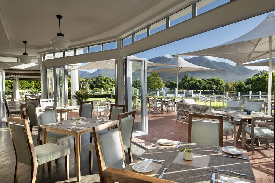 Ein modernes, elegantes Restaurant mit Holztischen und -stühlen, einem hellen, offenen Raum mit großen Fenstern, die einen Panoramablick auf üppiges Grün und Berge in der Ferne bieten.