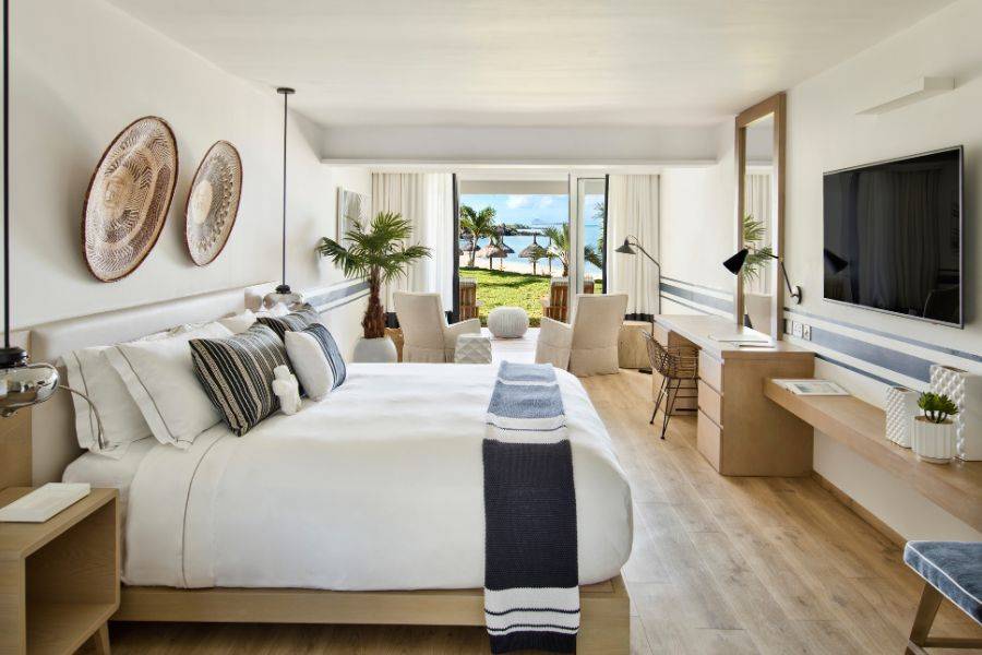 Modernes, helles Hotelzimmer mit gemütlichem Doppelbett, stilvollen Möbeln und Blick auf Palmen sowie das Meer durch eine geöffnete Terrassentür.