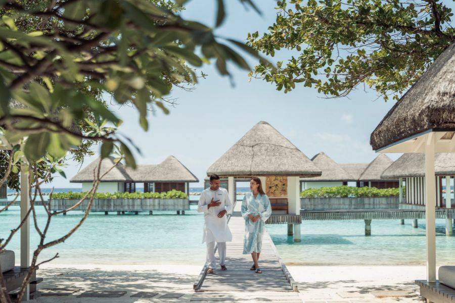  Eine Frau im Bademantel und ein Mitarbeiter im weißen Gewand spazieren entspannt über einen Holzsteg zwischen luxuriösen Wasserbungalows auf türkisfarbenem Meer.