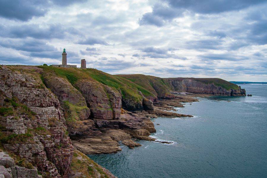 Blick auf die steilen Klippen des Cap Fréhel in der Bretagne mit dem markanten Leuchtturm und dramatisch bewölktem Himmel über dem Meer.
