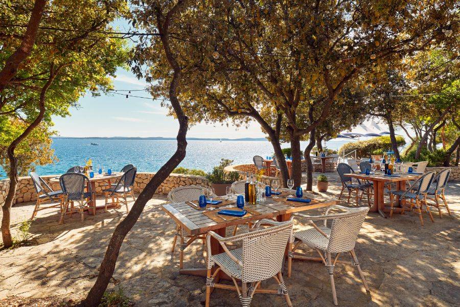 Gemütlich gedeckte Tische in einem schattigen Restaurant unter Bäumen, mit traumhaftem Blick auf das glitzernde Meer.