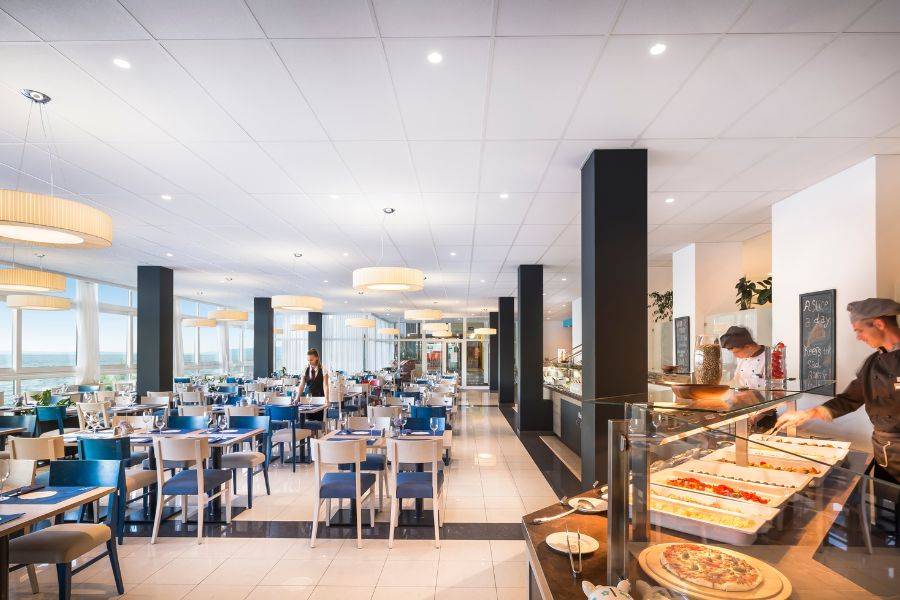 Helles Restaurant mit großen Fenstern, die einen Panoramablick auf das Meer und die Küste bieten, stilvollen Möbeln in Blau- und Naturtönen und einer eleganten Tischdekoration.