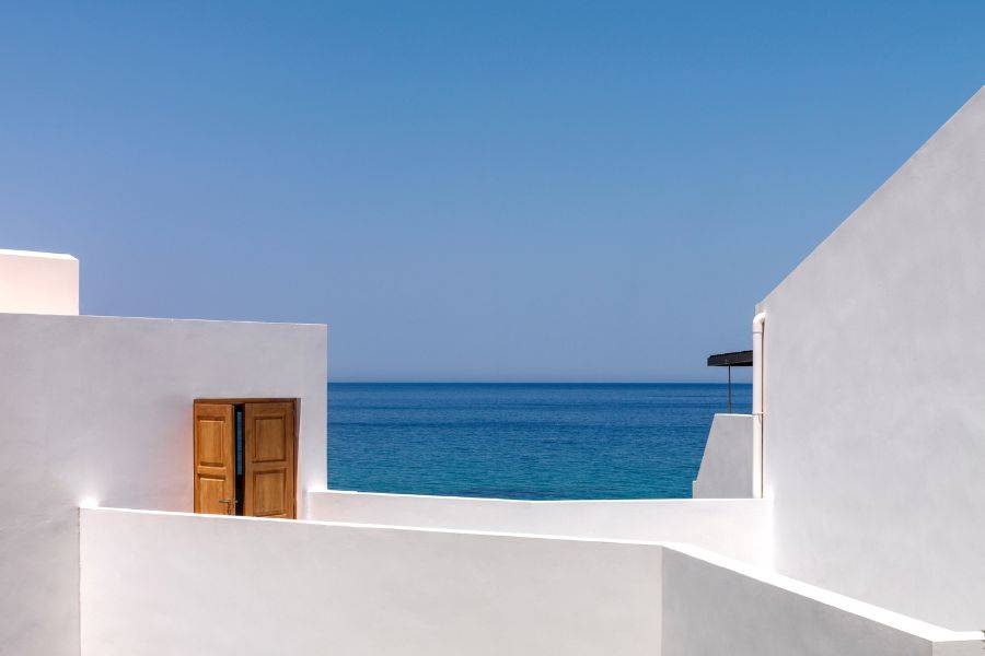 Weiße, minimalistische Gebäudestruktur mit hölzerner Tür, die sich zur offenen Aussicht auf das tiefblaue Meer unter klarem Himmel öffnet.