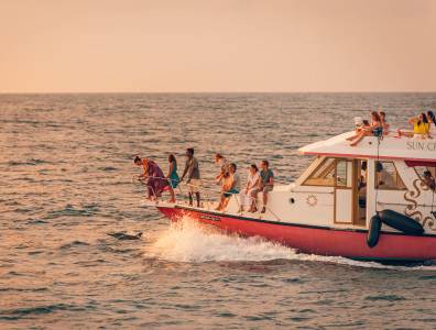 Personen auf einem kleinen Boot, welches am Wasser gleitet, während sich der Himmel orange färbt, weil die Sonne untergeht.