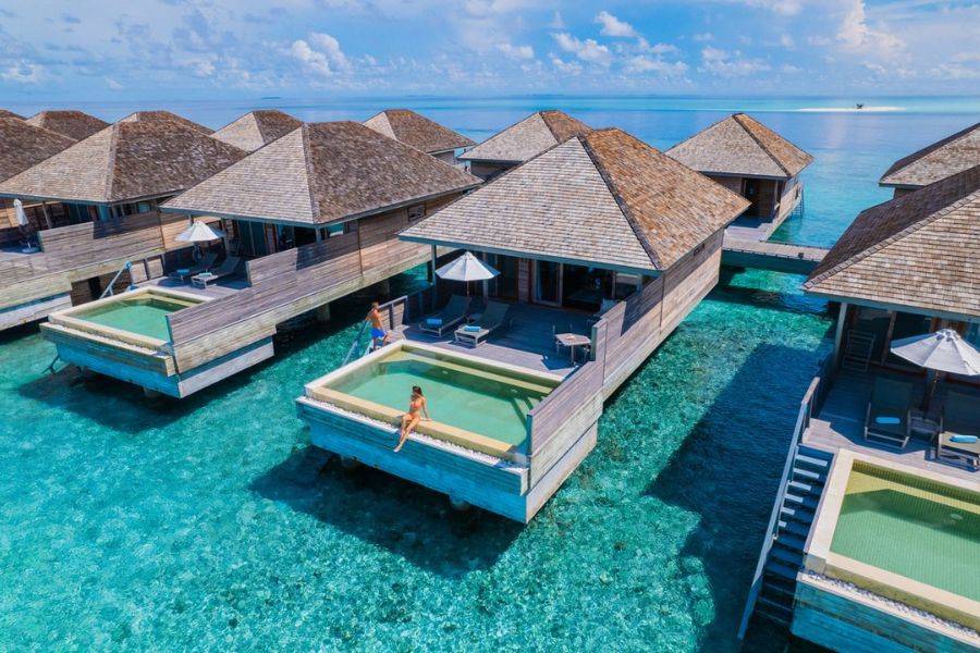 Eine traumhafte Wasserbungalow-Anlage auf den Malediven mit strohgedeckten Hütten, die über dem türkisblauen Wasser stehen. Der vordere Bungalow verfügt über eine private Terrasse mit Sonnenliegen, einem Sonnenschirm und einem kleinen Infinity-Pool, der nahtlos in das Meer überzugehen scheint.