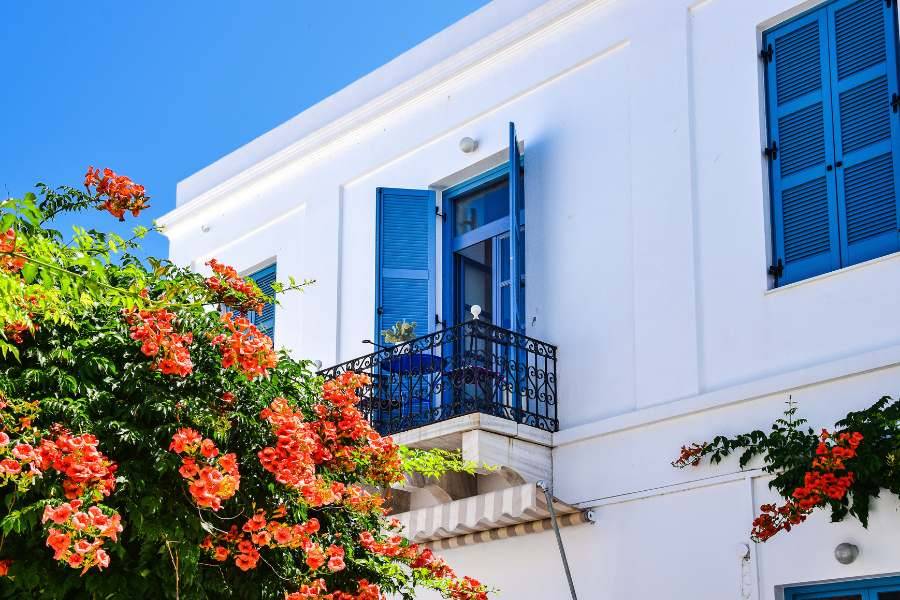 Weiße Fassade eines Hauses mit blauen Fensterläden und blühenden roten Blumen im Vordergrund.