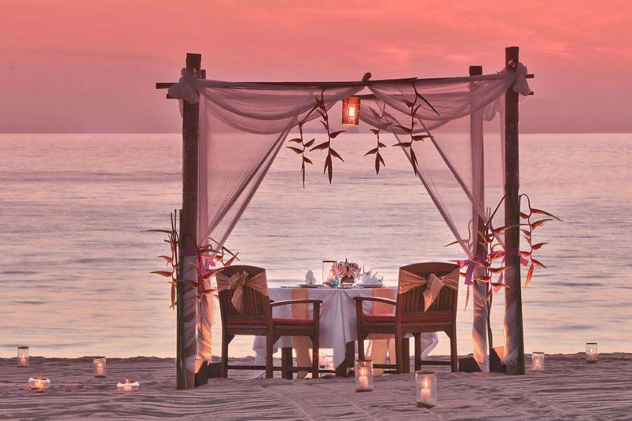 Romantisch dekorierter Tisch für zwei unter einem Pavillon aus Tüchern und Blüten direkt am Strand, bei Sonnenuntergang in warmem Licht.