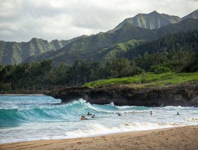 Wellen am naturbelassenen Strand der North Shore auf Oahu, Hawaii – grüne Berghänge, felsige Küste und Surfer im Wasser.