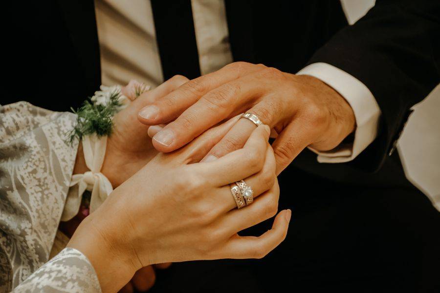 Nahaufnahme eines Paares, das sich bei der Hochzeit die Hände hält, beide tragen Eheringe und der Bräutigam trägt einen dunklen Anzug.