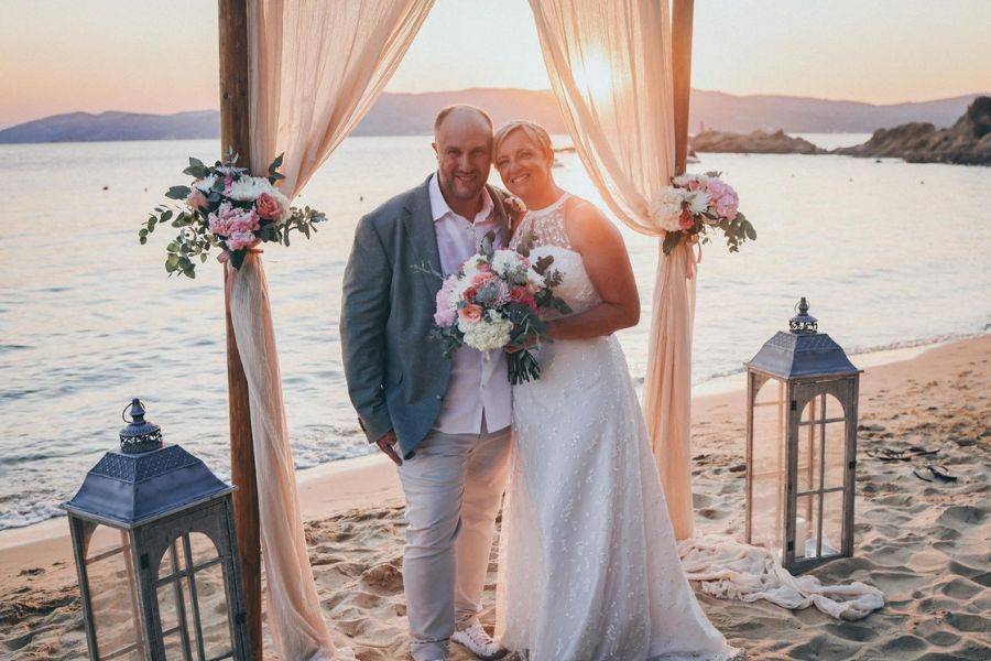 © privat Braut und Bräutigam posieren strahlend bei Sonnenuntergang unter einem mit Blumen geschmückten Hochzeitsbogen am Strand von Skiathos.