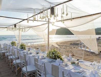 Festlich gedeckte lange Tafel am Strand unter einem transparenten, mit Blumen geschmückten Baldachin, mit Blick aufs Meer bei Sonnenuntergang.