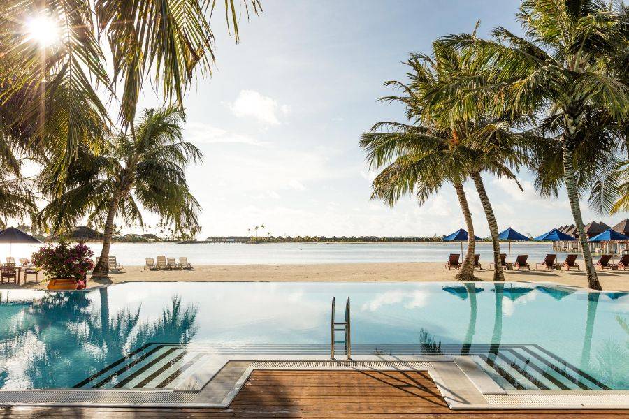 Moderner Poolbereich mit Einstiegstreppe und Meerblick, eingerahmt von Palmen und Sonnenliegen, mit Blick auf einen ruhigen, tropischen Strand.