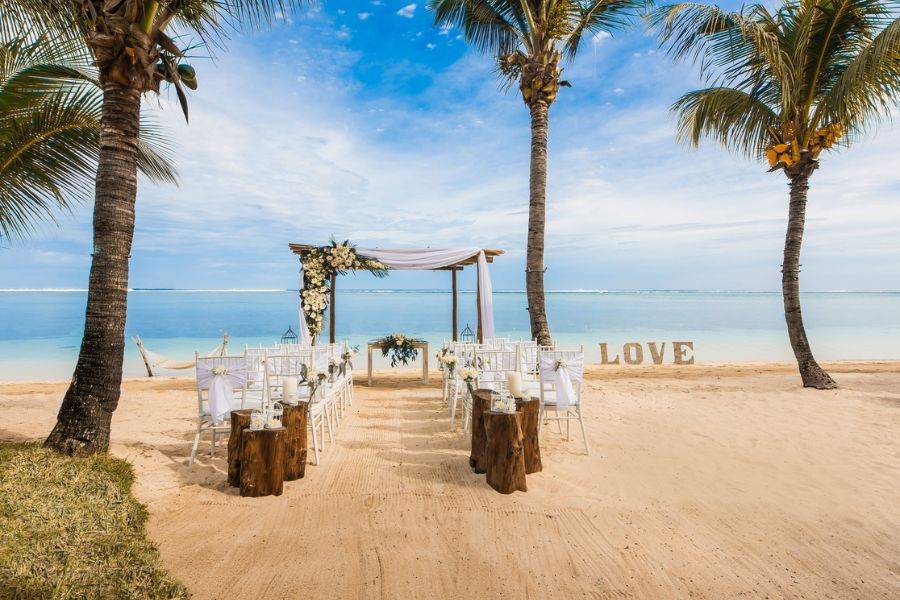 © LUX* Resorts Romantisch dekorierte Hochzeitskulisse am Strand zwischen Palmen mit weißem Pavillon, Stühlen und der Aufschrift „LOVE“ im Sand, vor türkisblauem Meer.