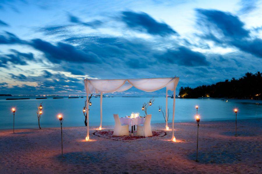 © Sheraton Maldives Full Moon Resort & Spa Ein gedeckter Tisch am Strand – Kerzenlicht bringt in der Abenddämmerung eine romantische Atmosphäre zum Leuchten.