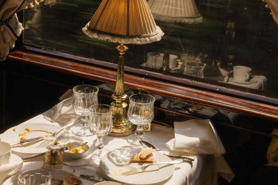 Ein elegant gedeckter Tisch in einem luxuriösen Zugrestaurant. Die Szene zeigt eine goldene Tischlampe mit einem klassischen Stoffschirm, mehrere leere Kristallgläser und teilweise verzehrte Speisen auf weißen Tellern.