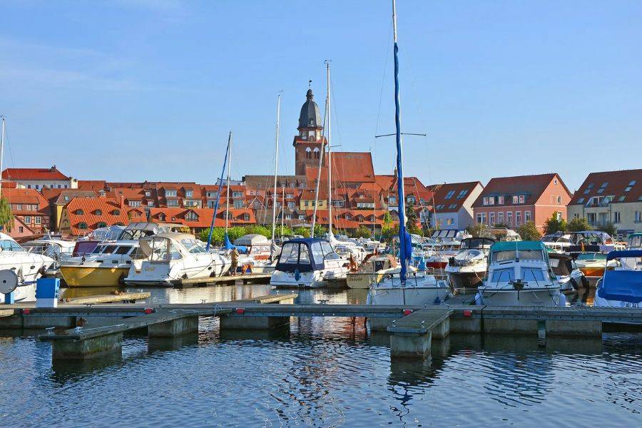 © Ruefa Yachthafen mit zahlreichen Motor- und Segelbooten im Vordergrund. Im Hintergrund ist eine malerische Stadt mit roten Ziegeldächern und einem markanten Kirchturm zu sehen.