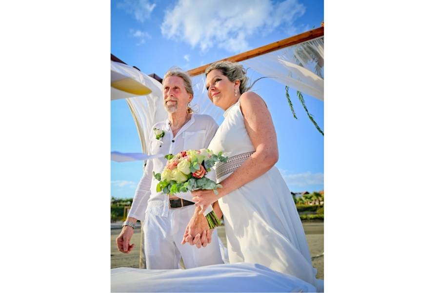 trandhochzeit auf Zypern: Brautpaar unter einem Hochzeitsbogen mit Blumenschmuck, in festlicher weißer Kleidung und mit Brautstrauß – Heiraten im Ausland bei Sonnenschein.
