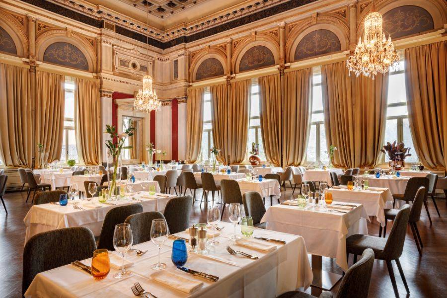 Elegantes Restaurant mit stilvoll gedeckten Tischen, großen Fenstern, cremefarbenen Vorhängen und weißen Blumen in hohen Vasen.
