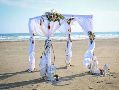 Romantischer Hochzeitsbogen am Strand von Zypern, dekoriert mit weißen Stoffbahnen, Blumenarrangements und Laternen – perfekte Kulisse für eine Strandhochzeit im Ausland.
