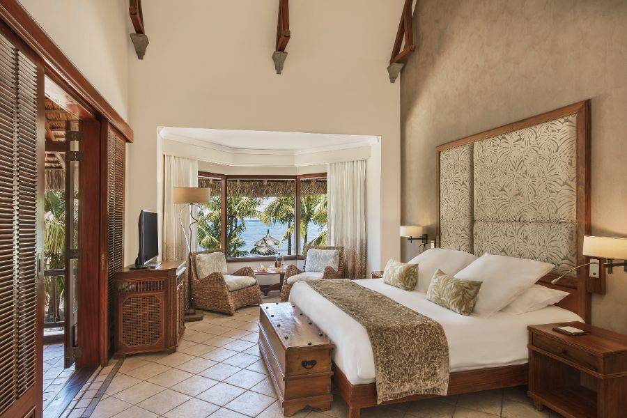 Luxuriöses Hotelzimmer mit Kingsize-Bett, eleganter Holzausstattung, Sitzecke mit Blick auf Palmen und das Meer durch große Fenster.