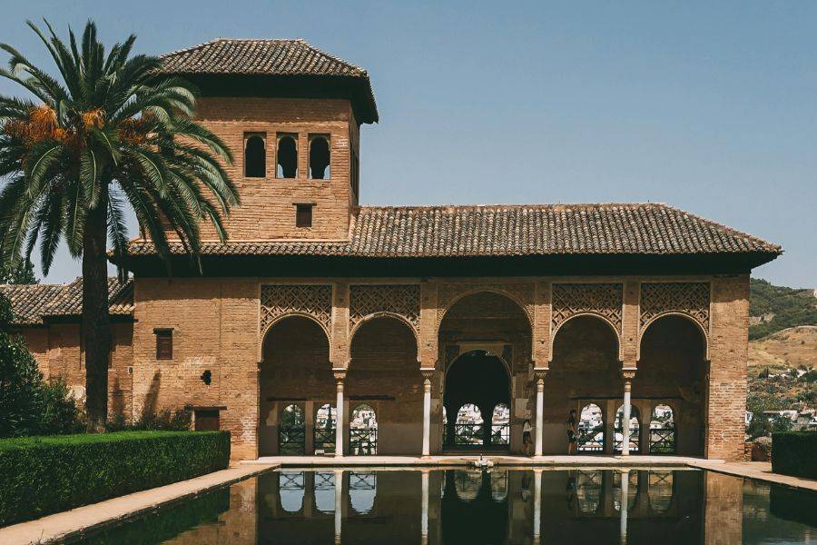Dieses Bild zeigt die Alhambra in Granada, eines der beeindruckendsten Beispiele maurischer Architektur in Spanien. Die filigranen Bögen, die warmen Erdtöne der Backsteine und die Spiegelung im Wasserbecken verleihen der Szene eine fast märchenhafte Atmosphäre.