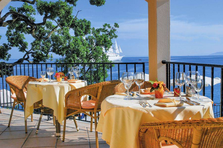 Charmante Terrasse eines Restaurants mit Blick auf das Meer, gedeckten Tischen mit gelben Tischdecken, Korbstühlen und einem vorbeifahrenden Segelschiff im Hintergrund.
