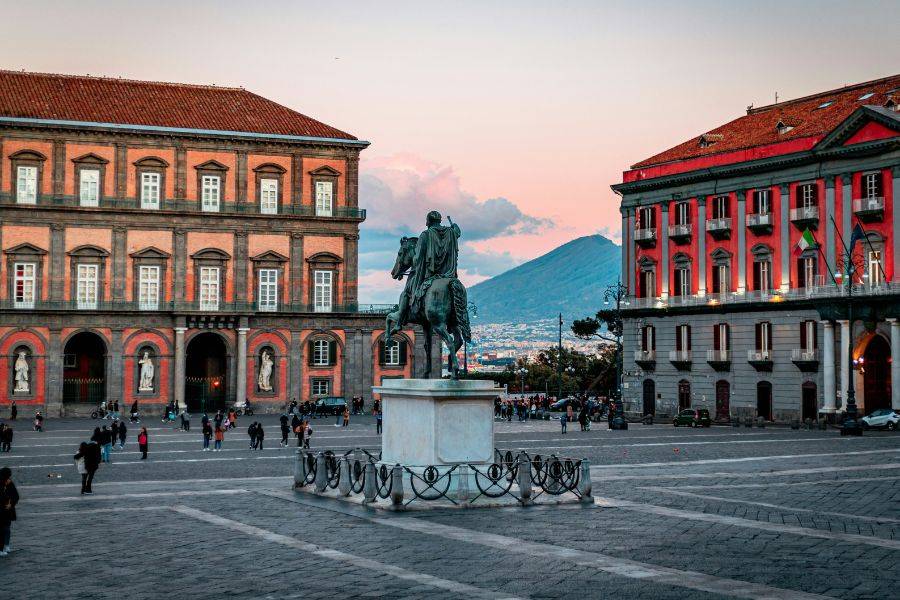 © Unsplash Platz in Neapel mit Reiterstatue im Vordergrund und dem Vesuv im Hintergrund, flankiert von historischen Gebäuden bei Sonnenuntergang.