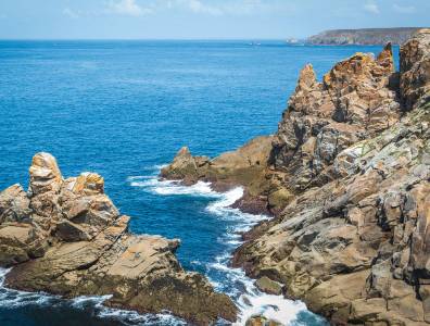 Wuchtige Felsen ragen an der bretonischen Küste aus dem blauen Atlantik, umspült von weißer Gischt unter blauem Himmel.