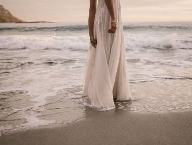 Eine Frau in einem fließenden weißen Kleid steht barfuß am Strand, während die Wellen sanft ihre Füße umspülen. Die Atmosphäre ist romantisch und verträumt.