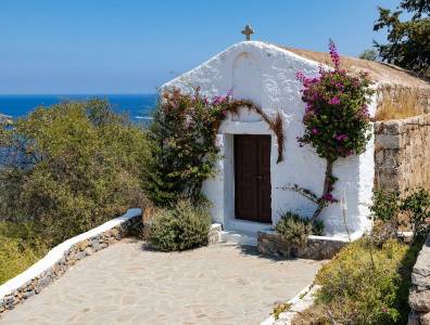 Eine kleine, traditionelle weiße Kapelle mit einer braunen Holztür, geschmückt mit rosa Bougainvillea-Blüten, thront auf einer Anhöhe mit Blick auf das blaue Meer.