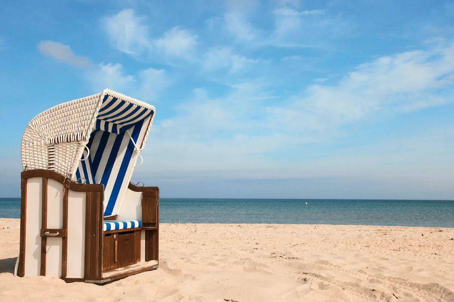 Ein einzelner, klassischer Strandkorb mit blau-weißen Streifen steht auf einem hellen Sandstrand, ausgerichtet zum ruhigen, blauen Meer.