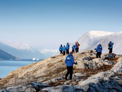 Expeditionsteilnehmer in blauen Jacken wandern entlang einer felsigen Küste in arktischer Landschaft mit Meer und Bergen