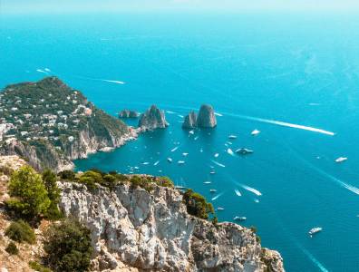 Blick auf die Insel Capri mit den berühmten Faraglioni-Felsen und zahlreichen Booten im tiefblauen Meer, aus luftiger Perspektive aufgenommen.