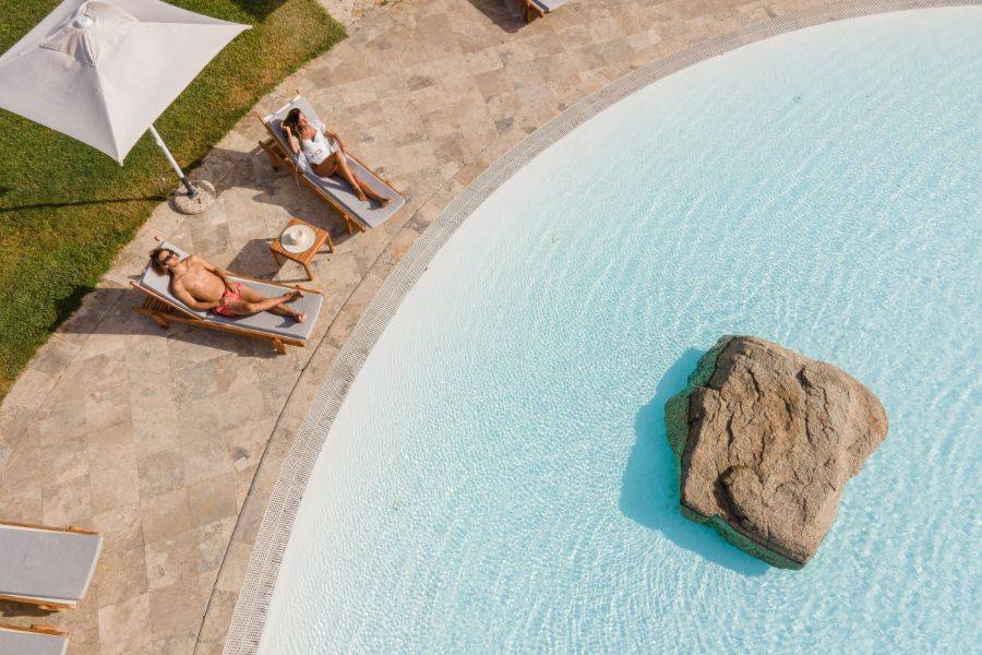 Ein Paar liegt entspannt auf Liegestühlen am Rand eines organisch geformten Pools mit Natursteinumrandung. Ein großer Felsen liegt im Wasser, ein Sonnenschirm spendet Schatten.