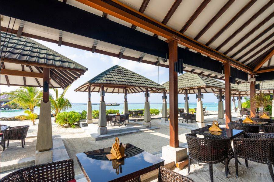 Offenes Strandrestaurant mit gedeckten Tischen unter Pavillons, umgeben von tropischen Pflanzen, mit Blick auf das Meer.